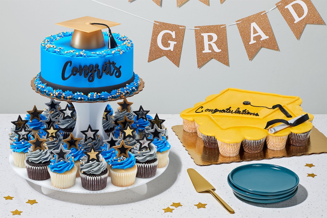 Graduation desserts with blue cake, cupcakes, yellow cupcake cake, mortarboard toppers, and “Grad” banner.