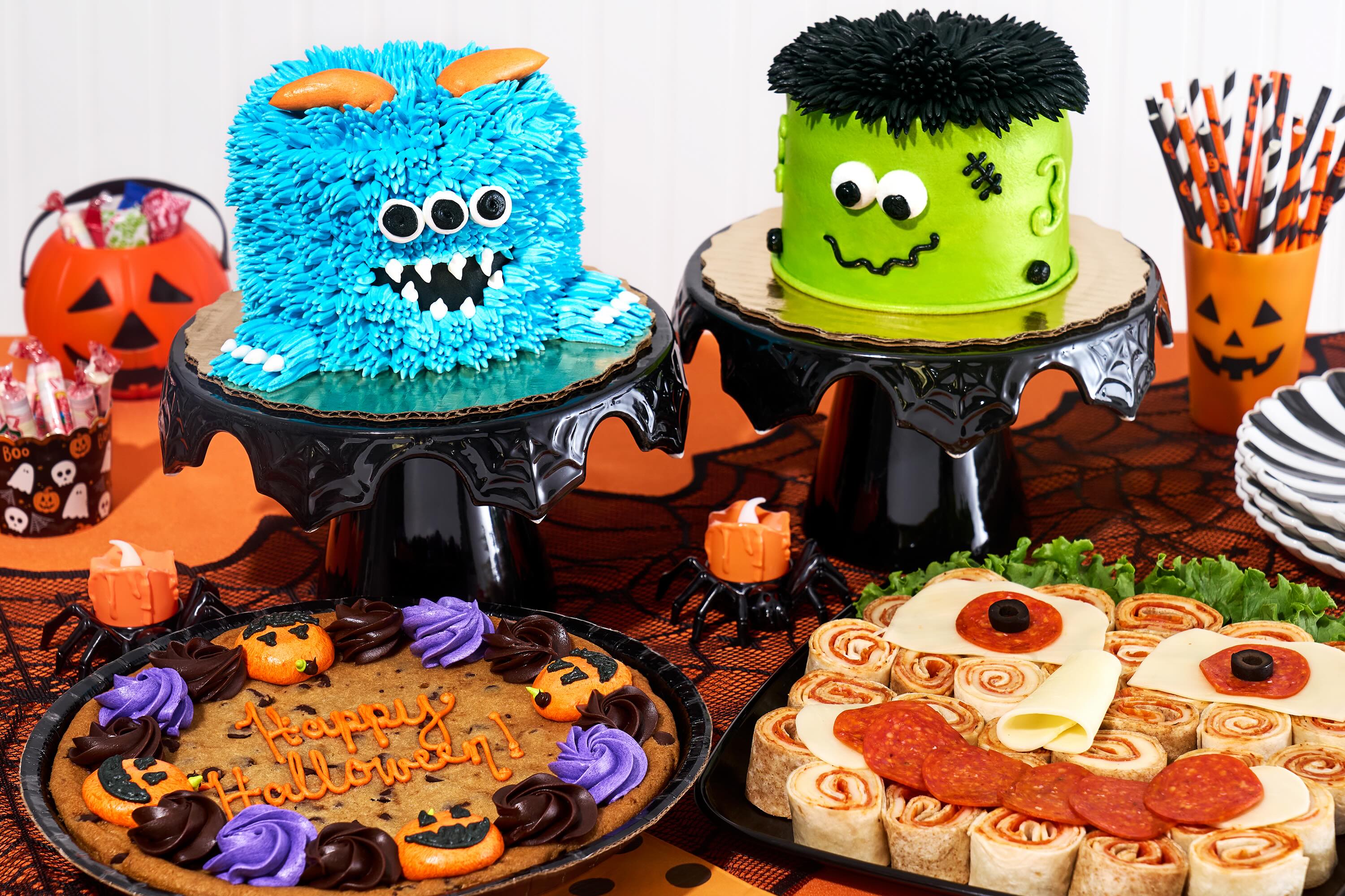 Halloween tablescape featuring a Pizza Roll-Up Platter, a monster mini cake next to a Frankenstein mini cake, and a large cookie decorated with the words “Happy Halloween.”