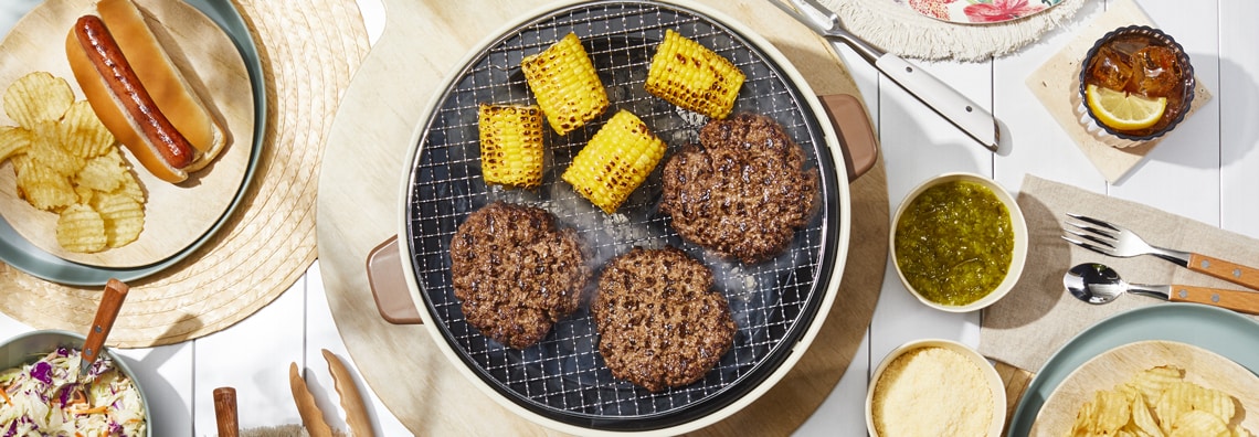 A summer barbecue spread with grilled burgers, corn, hot dogs, chips, coleslaw, relish, and iced tea on a table.