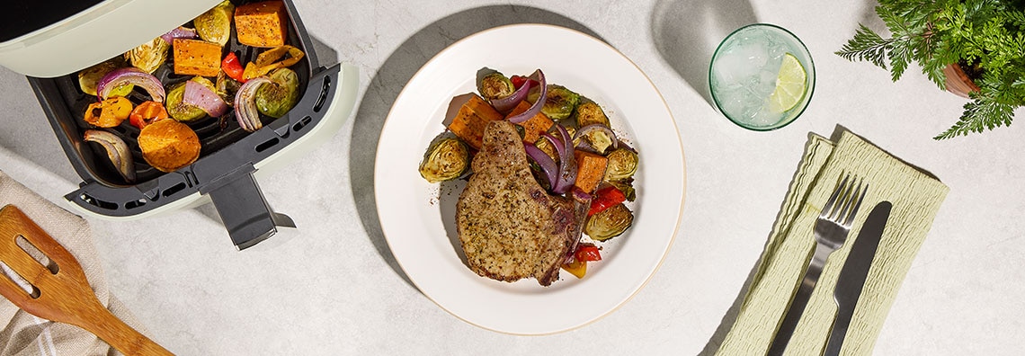 A plate with a cooked pork chop and roasted vegetables next to an air fryer basket filled with more roasted vegetables.