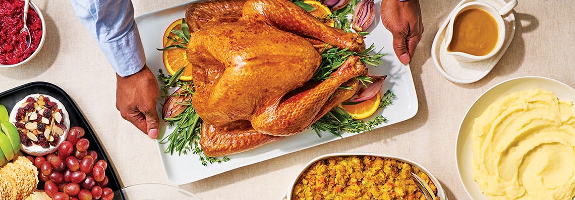 A Thanksgiving feast with a roasted turkey, mashed potatoes, stuffing, gravy, and a platter of grapes and cheese on a table.