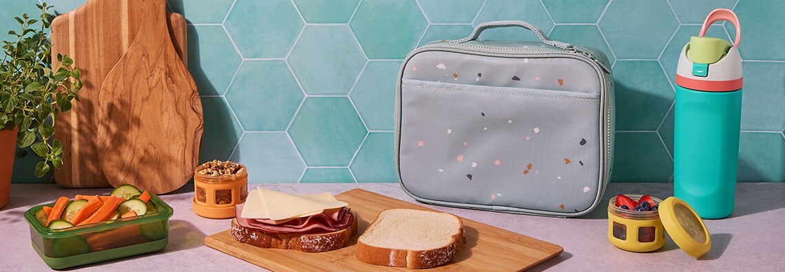 Lunch setup with a sandwich, veggies, nuts, and berries in containers, a water bottle, and a lunch bag on a counter with a teal tile background.
