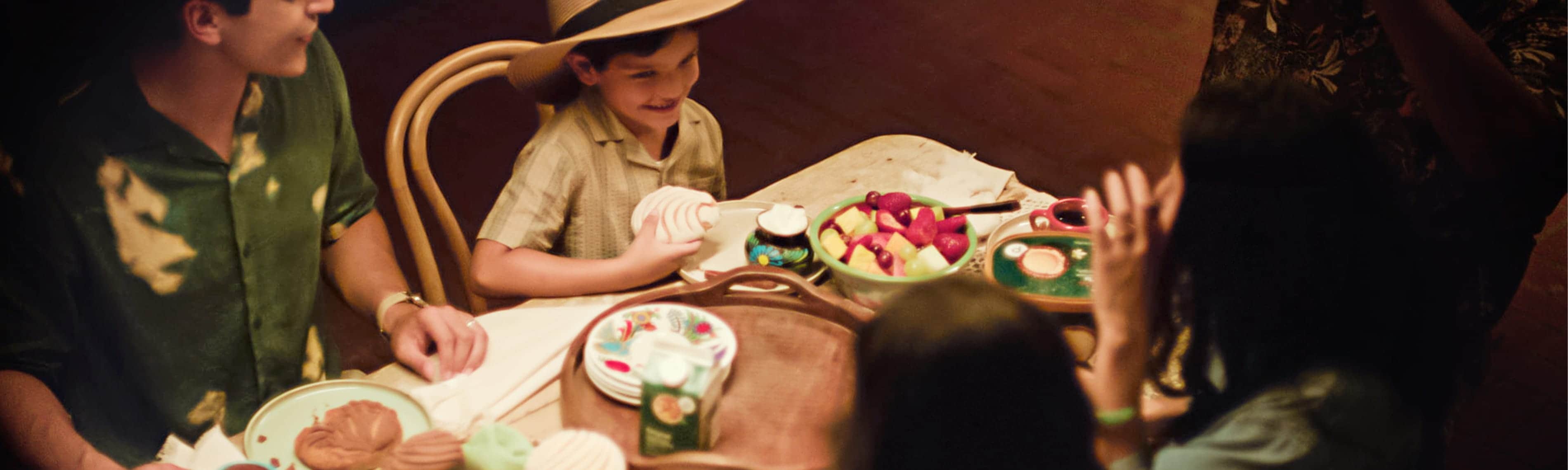 Family gathered around a dining table with fresh fruit, pastries, and snacks during a shared meal in a warm indoor setting.