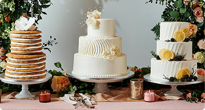 Three-tier white wedding cake with floral details centered between stacked Baked with Love cake and Blackberry Lemon Bliss cake on elegant dessert table.