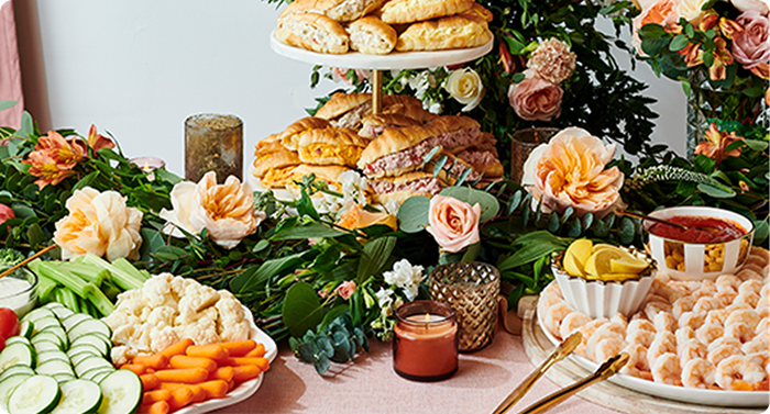 Wedding catering display with sandwiches, pastries, shrimp platter, fresh vegetables, and elegant floral arrangements on reception table.