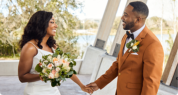 Couple holding hands during outdoor wedding, one holding bouquet with blush roses, standing by lakeside venue structure.