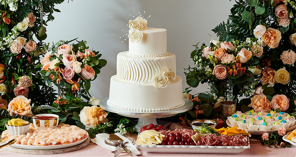 Three-tier white wedding cake with floral accents on elegant dessert table surrounded by blush roses, appetizers, and cupcakes at reception.