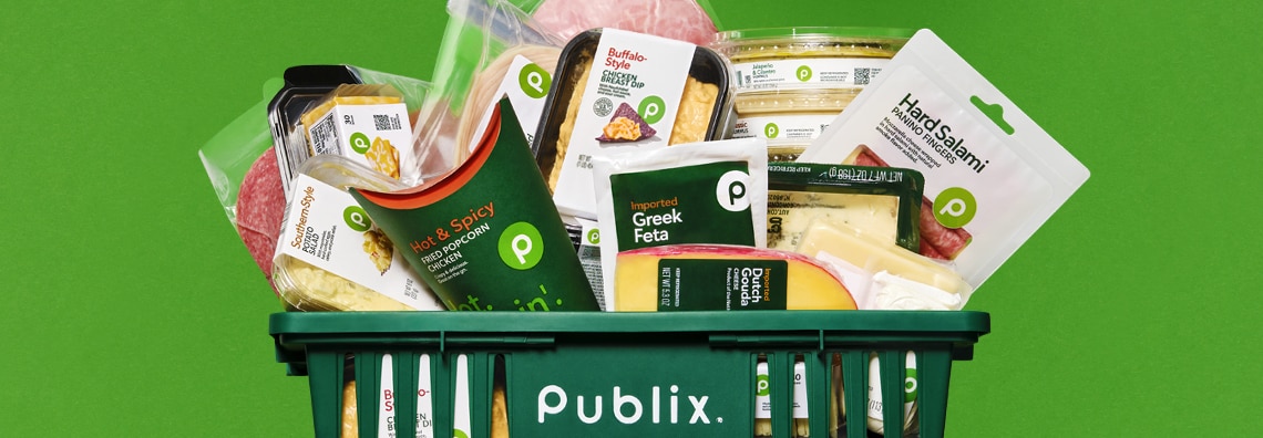 Publix shopping basket filled with various Publix brand deli and cheese products, including Greek feta, Dutch gouda, hard salami, hot and spicy fried popcorn chicken, and buffalo-style chicken dip, against a bright green background.