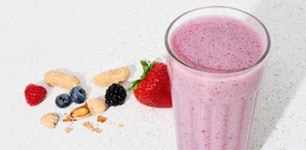 A glass of pink smoothie surrounded by fresh berries and peanuts on a white surface.
