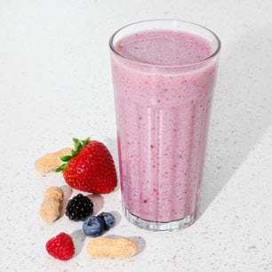 A glass of pink smoothie surrounded by fresh berries and peanuts on a white surface.