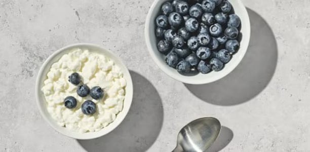 A bowl of fresh blueberries next to a bowl of cottage cheese topped with five blueberries. Both bowls are on a marble countertop, with a spoon shown partially in the frame.