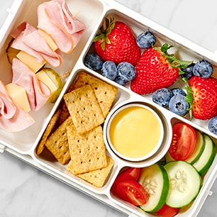 Balanced lunchbox with turkey roll-ups, cheese, strawberries, blueberries, crackers, veggie slices, and a creamy dipping sauce.