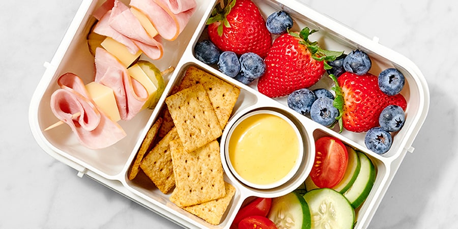 Balanced lunchbox with turkey roll-ups, cheese, strawberries, blueberries, crackers, veggie slices, and a creamy dipping sauce.