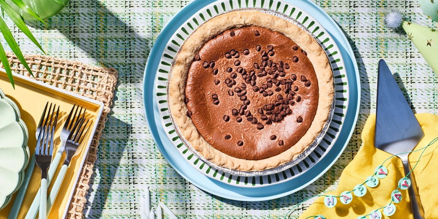 A chocolate pie topped with chocolate chips on a decorative plate, surrounded by forks, a pie server, and a Happy Birthday banner.