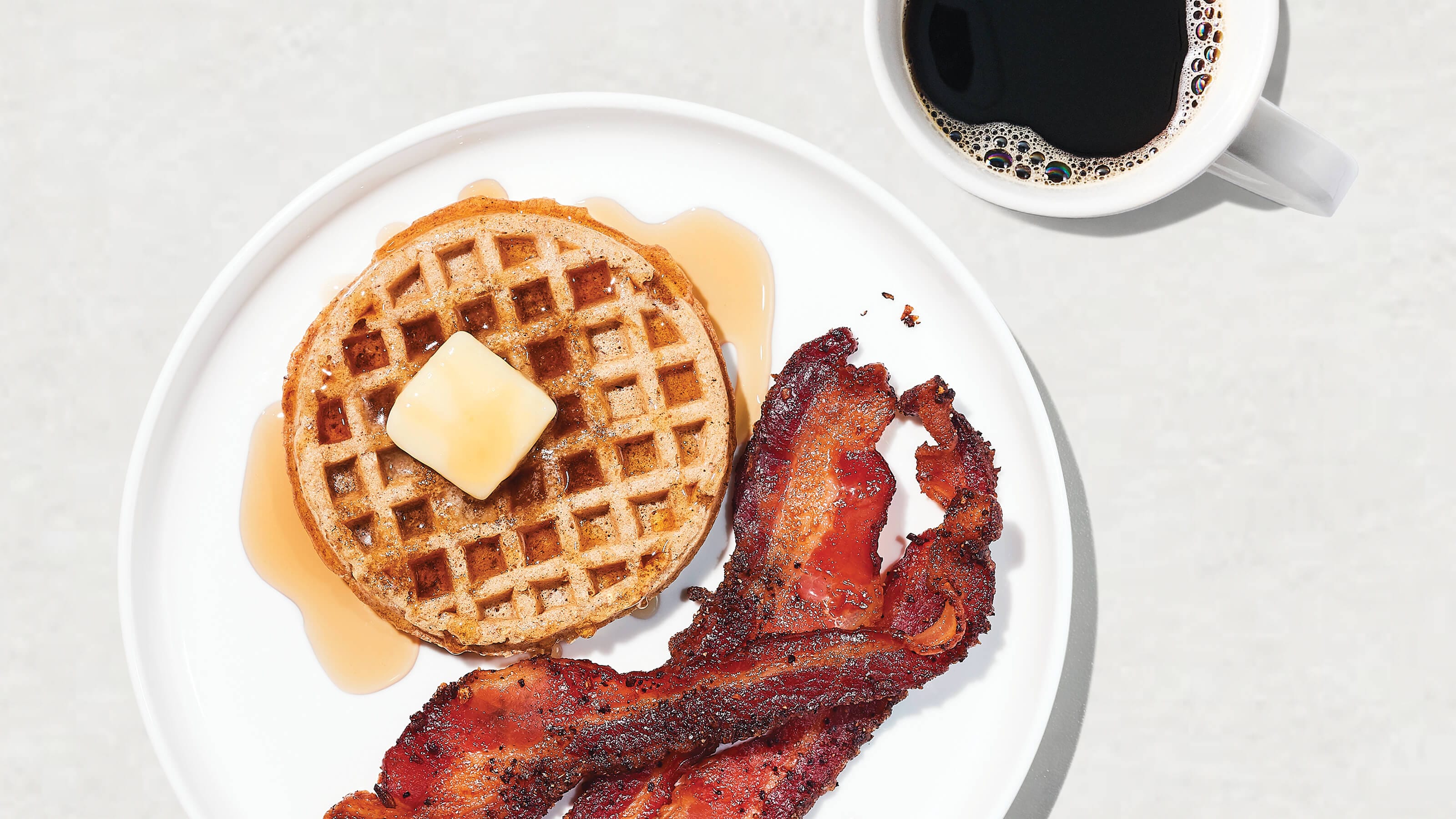 A plate with a waffle topped with butter and syrup, crispy bacon strips, and a cup of black coffee on the side.