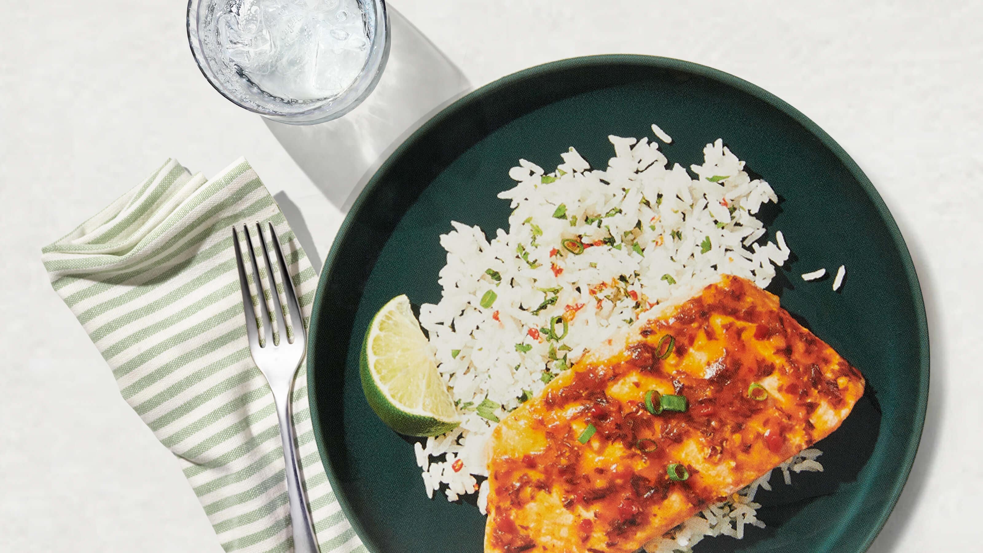 Plate of glazed salmon with white rice, garnished with green onions and lime wedge, next to a striped napkin and glass of ice water.