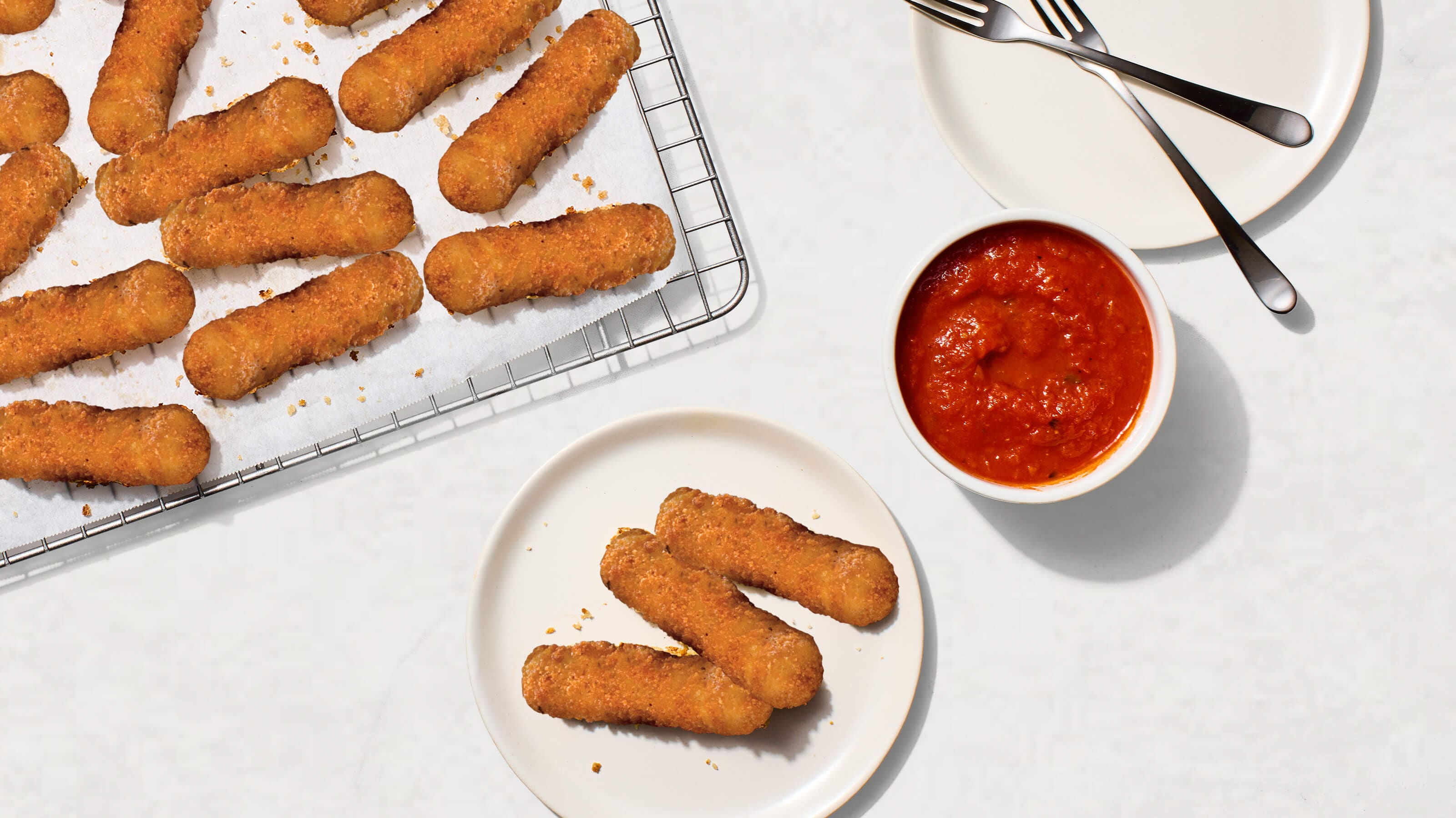 Mozzarella sticks on a cooling rack and plate, served with marinara sauce in a bowl, alongside forks on a white table.