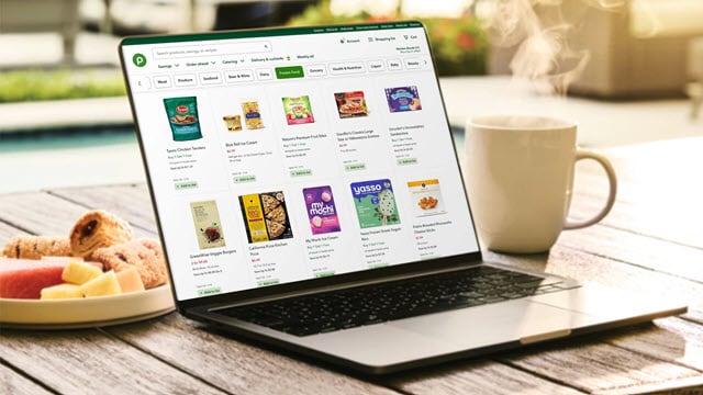A laptop displaying a Publix online shopping page with frozen food items, placed on a table next to a coffee mug and a plate of snacks.