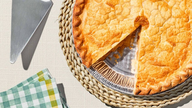 A golden-brown pie with a slice removed, placed on a woven mat, alongside a pie server and green checkered napkins.