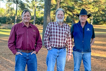 Three men are standing outdoors at Prestage Farms surrounded by trees.
