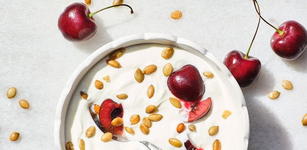 A bowl of yogurt topped with cherries and seeds, surrounded by fresh cherries on a light surface
