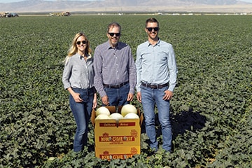 Three people standing in a large crop field behind a crate of cantaloupes on Turlock Fruit Company farm.