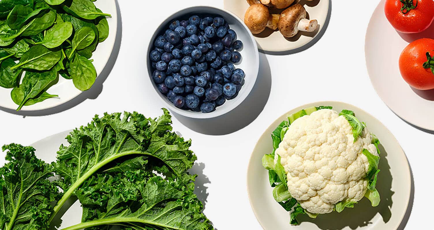 Overhead view of fresh vegetables and fruit—spinach, kale, cauliflower, blueberries, mushrooms, and tomatoes—arranged in bowls on a white background.