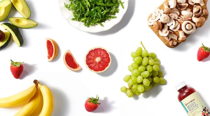 Overhead arrangement of fresh organic produce on a white background, including avocados, bananas, strawberries, green grapes, grapefruit halves, mushrooms, leafy greens, and a bottled juice.
