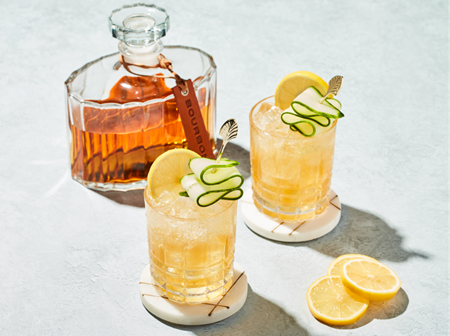 A "Bourbon" decanter with two cocktail glasses garnished with lemon and cucumber, set on coasters, with lemon slices nearby on a lit surface.