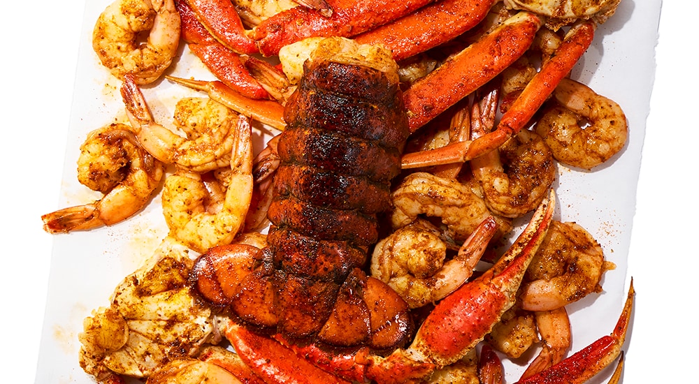 A platter of steamed and seasoned seafood including shrimp, crab legs, and a lobster tail, arranged on a white surface.