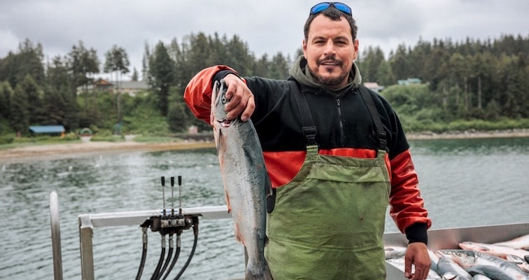 A fisherman holding a fresh caught fish infront of a lake