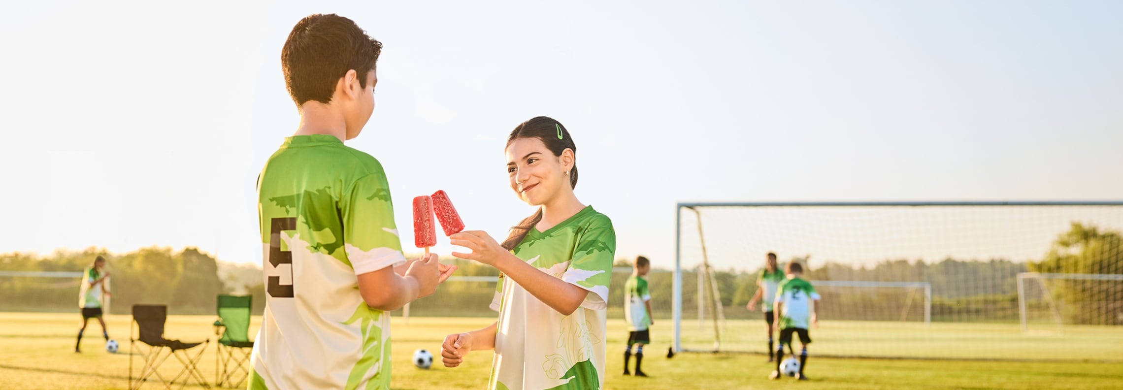 Two youth soccer players share ice pops on the sidelines while teammates practice on a soccer field in the background.