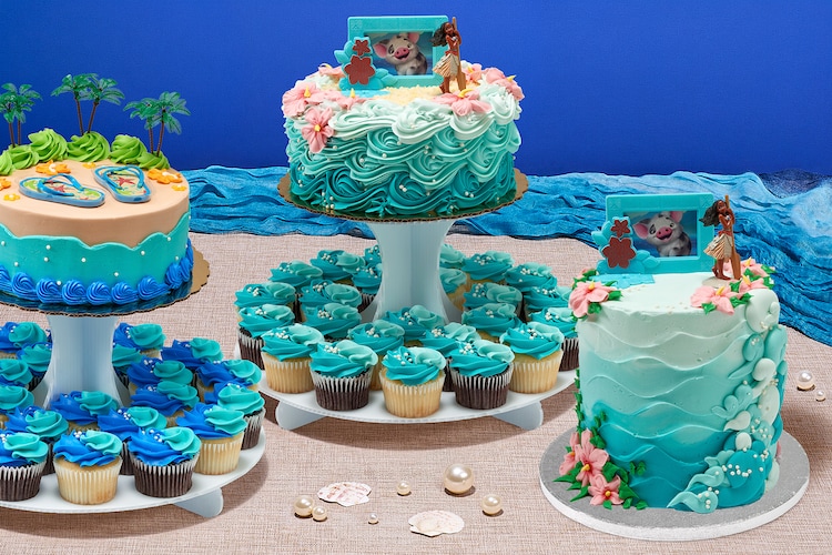 Moana and summer-themed cakes on a sand-colored surface with seashells and blue fabric waves.