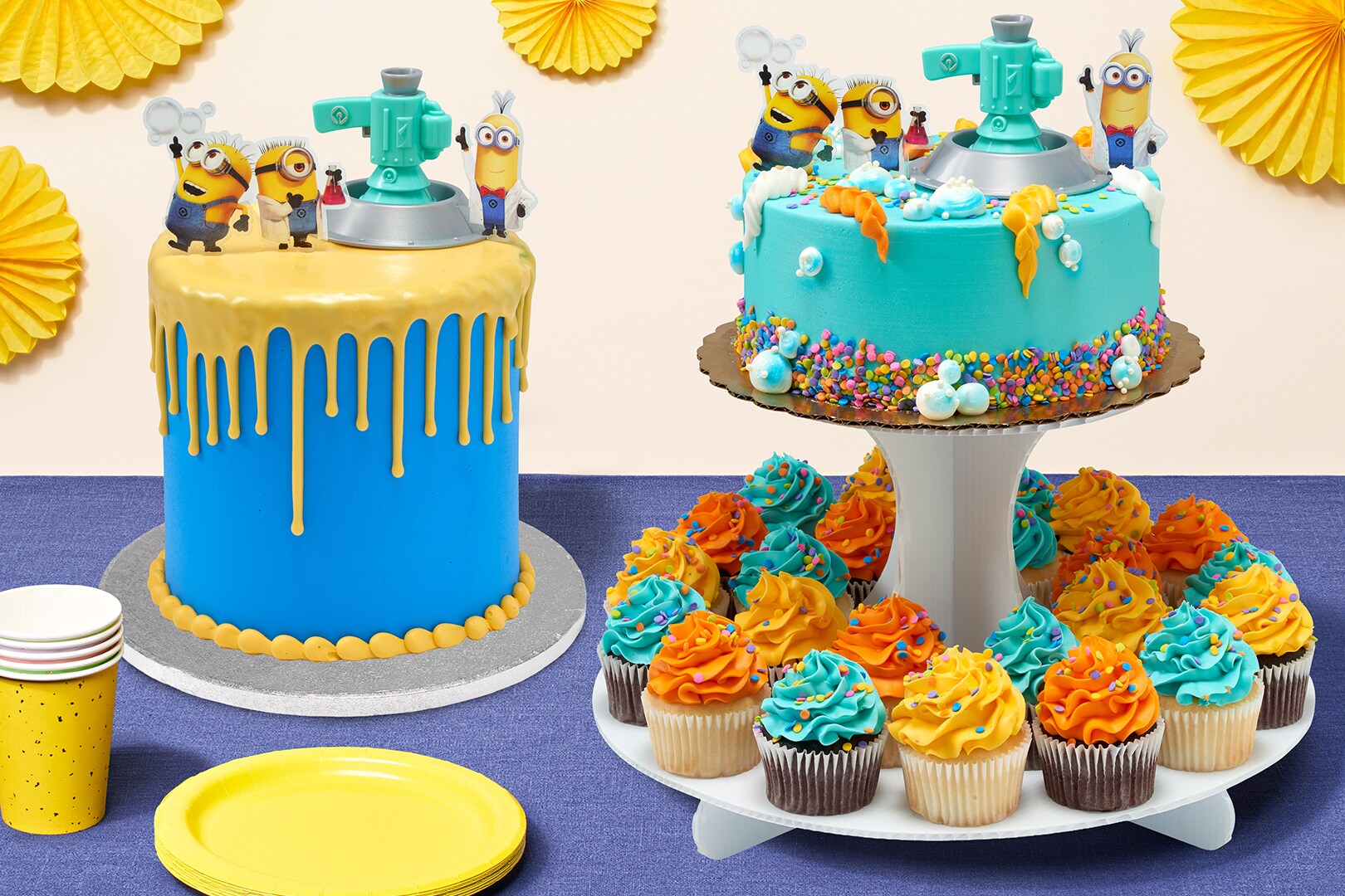 Minions Celebration Towering Tier Cake and cake-cupcake combo on a blue surface with yellow plates, cups, and fan decorations.  Minions Franchise c Universal City Studios LLC. All rights reserved